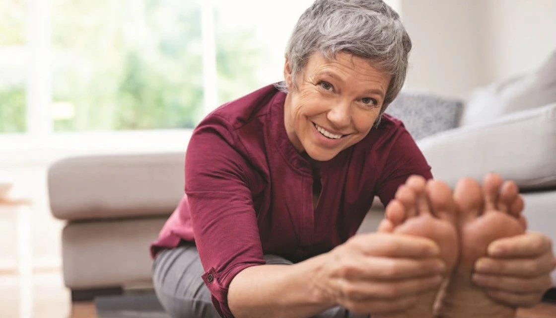 Stock image of a senior stretching and reaching for their toes. The smiling senior has short grey hair and is wearing a burgundy shirt and grey pants.