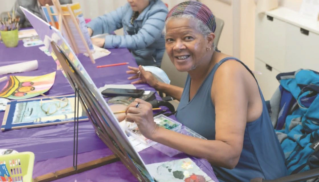 Image used for the 'Stay active and socially connected at your On Lok PACE center' blog post. The participant is painting and smiling at the camera.
